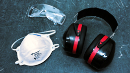 A clear safety goggles, a white mask, and black ear protection with red accents on a dark surface.
