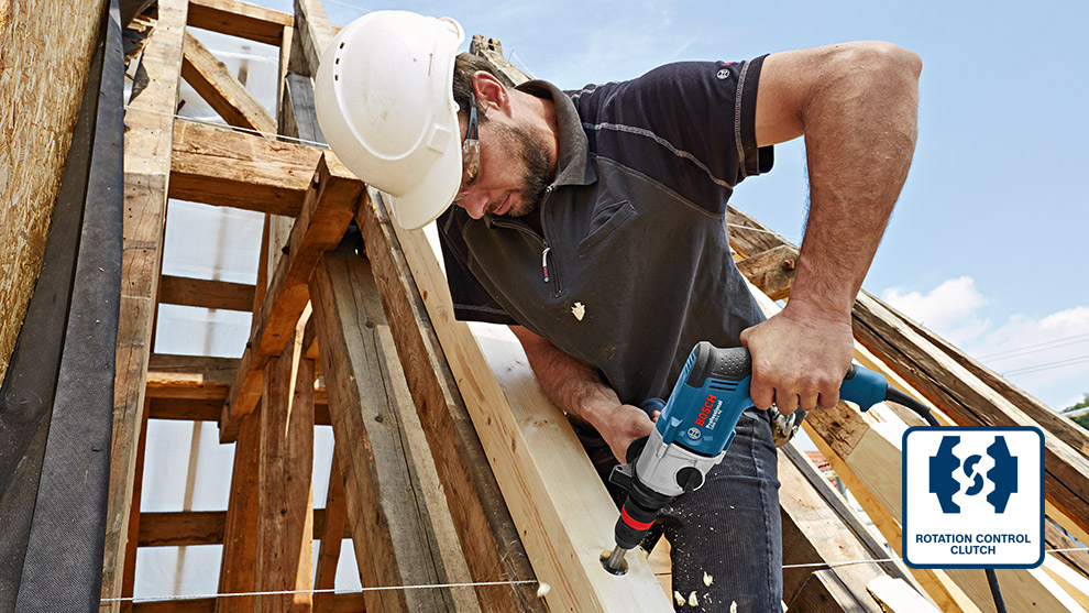 A man in a helmet uses a Bosch drill on wooden beams outdoors.