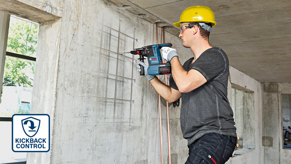 A worker in a yellow helmet uses a Bosch drill on a concrete wall indoors.